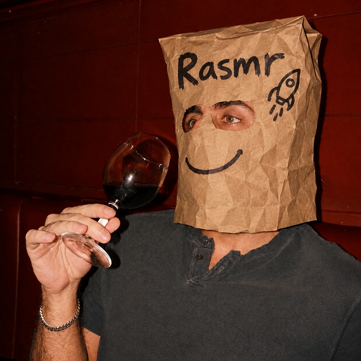A man wearing a paper bag with "Rasmr" written in marker, holding a glass of red wine